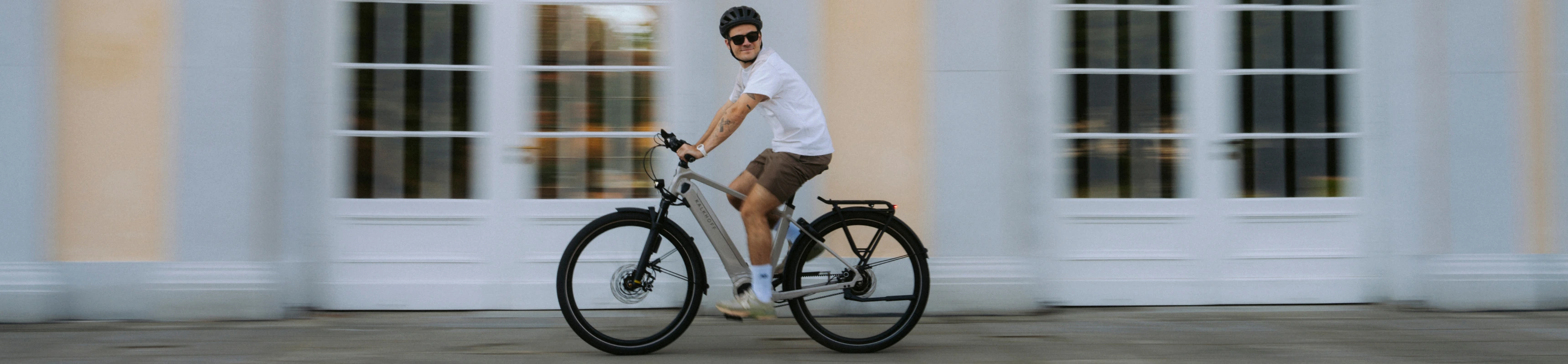 Mann in kurzer Hose fährt mit seinem beigen Kalkhoff E-Bike durch die Stadt an einem hellen Gebäude entlang
