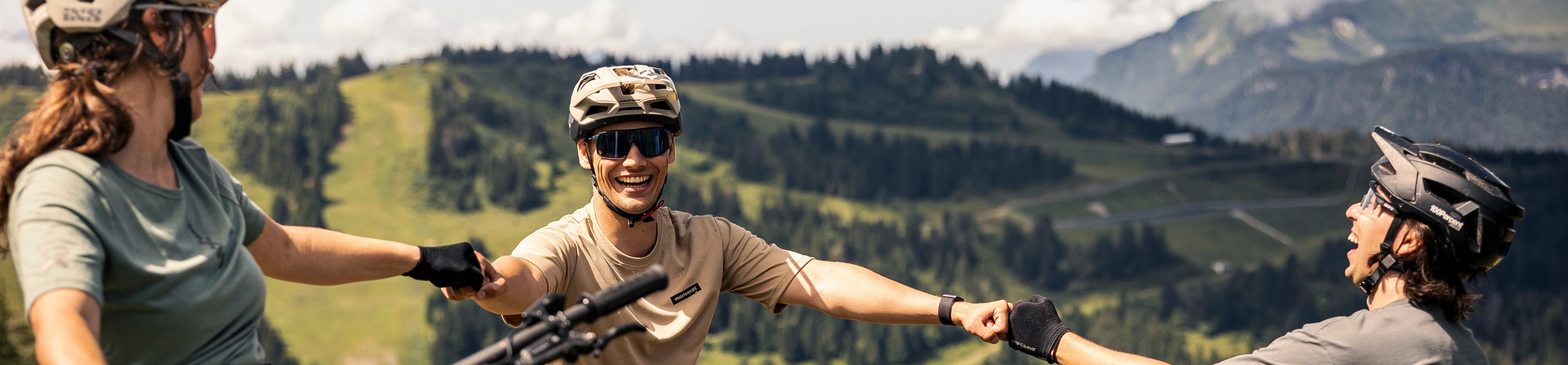 Drei Mountainbiker geben sich die Faust und lachen im Hintergrund sieht man grüne Hügel